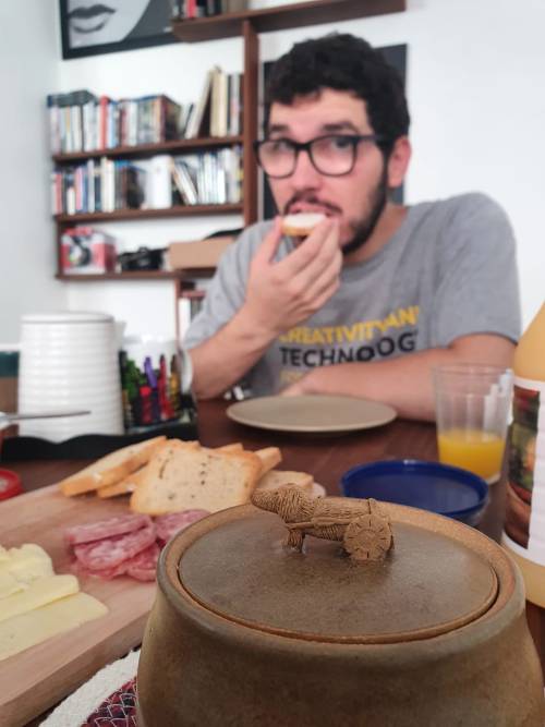 Uma foto em ambiente interno mostrando um homem de óculos e barba, usando uma camiseta cinza, comendo algo durante uma refeição. Na mesa há frios, queijos e biscoitos. Em primeiro plano, há um recipiente marrom com tampa decorada com um pequeno ornamento. Ao fundo, vê-se uma estante de madeira com livros e um quadro na parede.