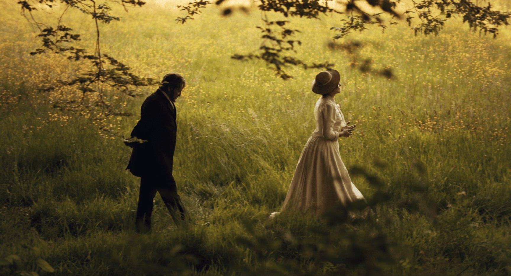 Fotografia artística em tons dourados de duas pessoas em um campo alto de grama e flores silvestres. Uma veste um vestido branco vitoriano e chapéu, a outra usa trajes escuros formais. Ambas caminham separadamente em meio à luz do sol da tarde que banha a cena.